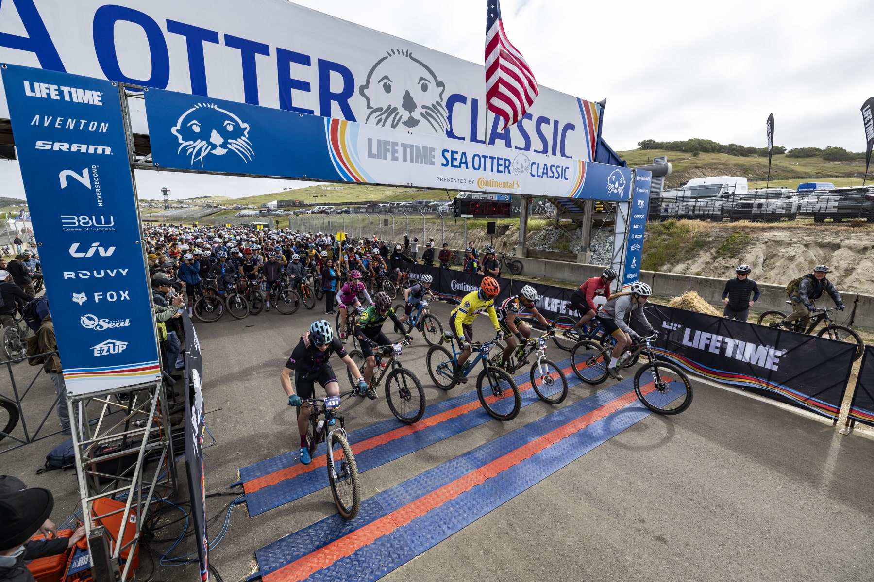 Salida de una carrera de BTT en el Sea Otter Classic en Monterey, California con numerosos ciclistas iniciando la prueba bajo el arco oficial del evento de ciclismo.